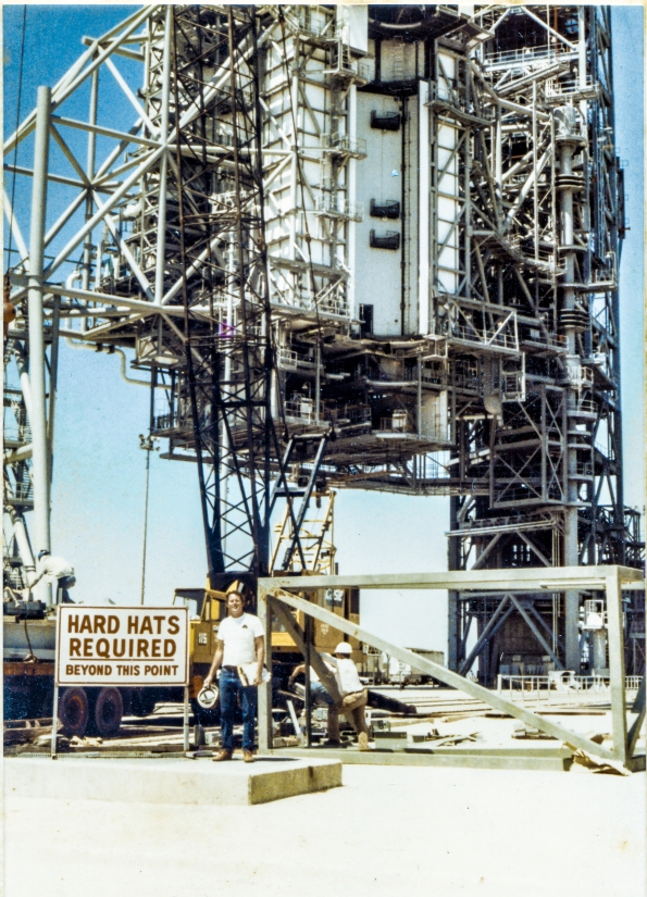 Image 055. Our earliest look at James MacLaren at Space Shuttle Launch Complex 39-B, Kennedy Space Center, Florida, after returning to the jobsite following his layoff from Sheffield Steel, and his subsequent hiring by Ivey Steel. In the far distance, the RSS and FSS loom into the sky, overflowing the frame, sensibly-complete in general appearance, but far from complete for the purposes which they were designed and constructed for, servicing the Space Shuttle, prior to launch. In the near distance, the yellow 90-ton P&H crane is being used to offload more steel from the back of a flatbed semitrailer. Union Ironworkers from Local 808 work the load from on top of the trailer and on the ground next to it, while the white-shirted truck driver lounges against the lopped-off tube steel top of the East Stair Tower. In his left hand, MacLaren grips a clipboard holding the sheets of the shipping list that came with the load of iron, with a yellow notepad behind it. In his right hand, he holds his hard hat, and if you look very close, you can also see a yellow highlighter in that hand for marking up the shipping list indicating items which have been checked and verified as being included in this delivery. On his right hip, a 25-foot tape measure that stayed with him wherever he went on the Pad Deck or on the Towers. Another day on the job. Photo by James MacLaren. Image 055. Our earliest look at James MacLaren at Space Shuttle Launch Complex 39-B, Kennedy Space Center, Florida, after returning to the jobsite following his layoff from Sheffield Steel, and his subsequent hiring by Ivey Steel. In the far distance, the RSS and FSS loom into the sky, overflowing the frame, sensibly-complete in general appearance, but far from complete for the purposes which they were designed and constructed for, servicing the Space Shuttle, prior to launch. In the near distance, the yellow 90-ton P&H crane is being used to offload more steel from the back of a flatbed semitrailer. Union Ironworkers from Local 808 work the load from on top of the trailer and on the ground next to it, while the white-shirted truck driver lounges against the lopped-off tube steel top of the East Stair Tower. In his left hand, MacLaren grips a clipboard holding the sheets of the shipping list that came with the load of iron, with a yellow notepad behind it. In his right hand, he holds his hard hat, and if you look very close, you can also see a yellow highlighter in that hand for marking up the shipping list indicating items which have been checked and verified as being included in this delivery. On his right hip, a 25-foot tape measure that stayed with him wherever he went on the Pad Deck or on the Towers. Another day on the job. Photo by James MacLaren.